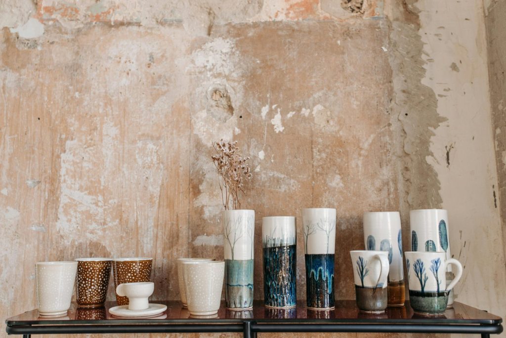 A collection of handmade decorated ceramic cups on a rustic wooden table against a textured wall.